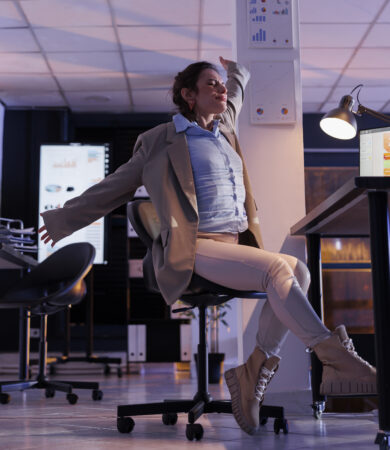 Femme d’affaires s’étirant devant son bureau dans un environnement professionnel moderne, symbolisant la fatigue, la gestion du temps ou le besoin d’organisation.