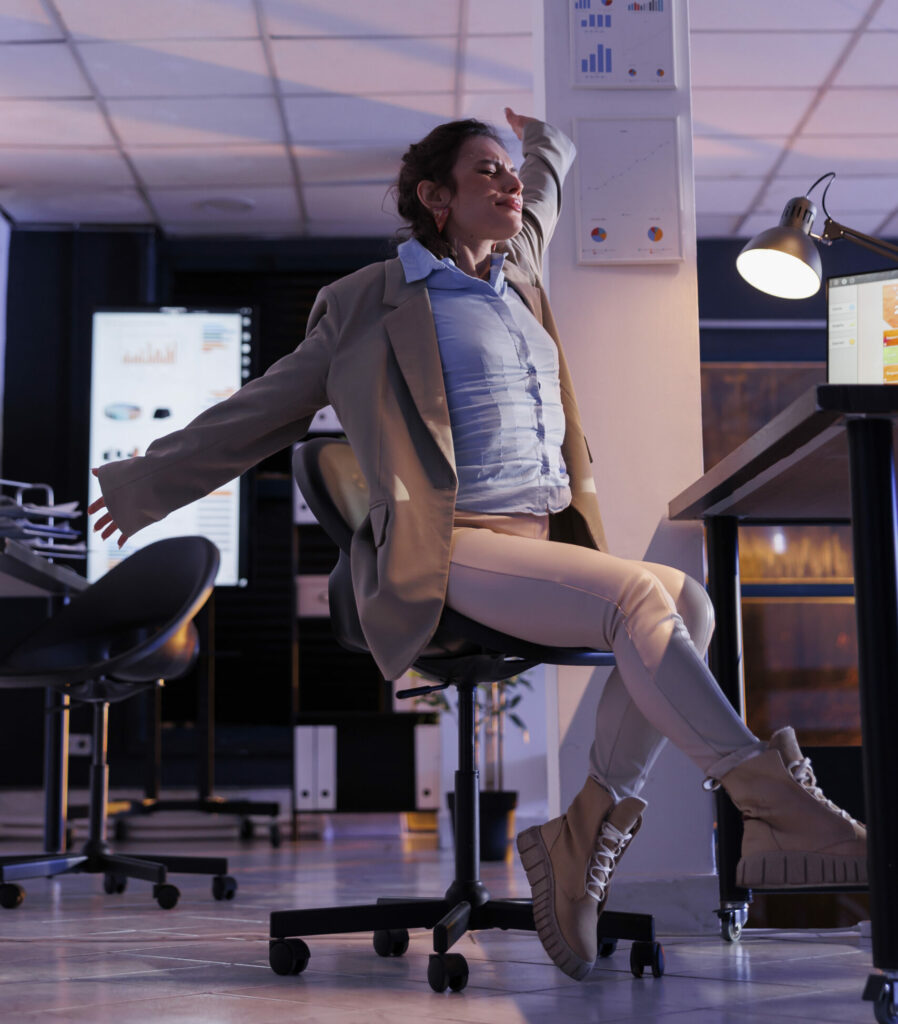 Femme d’affaires s’étirant devant son bureau dans un environnement professionnel moderne, symbolisant la fatigue, la gestion du temps ou le besoin d’organisation.